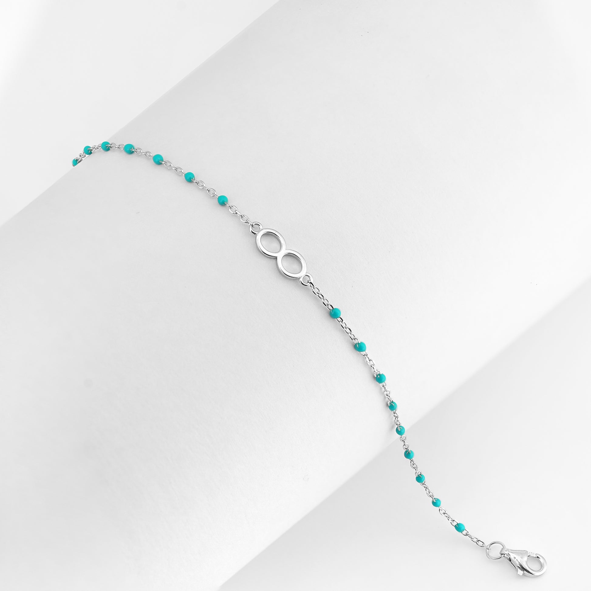A delicate silver bracelet with turquoise beads and an infinity symbol shown on a white background.

