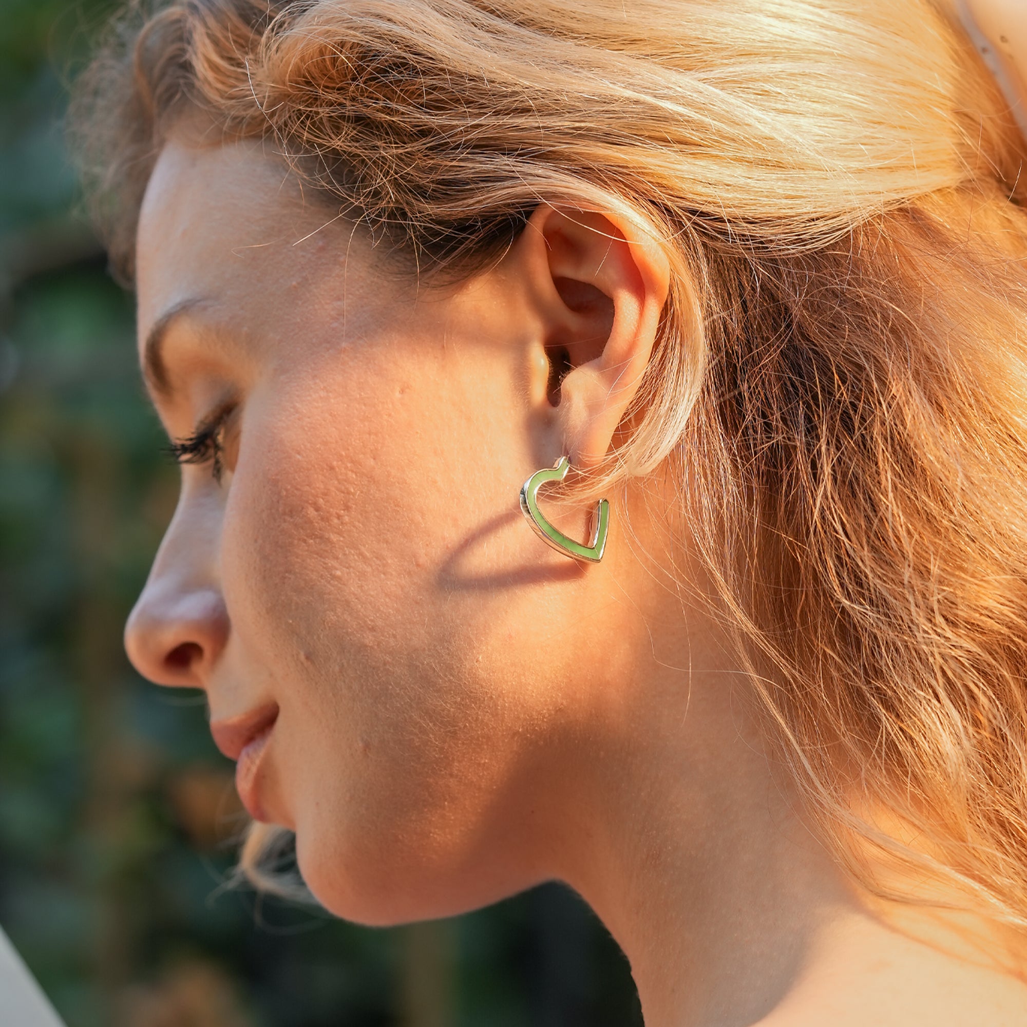 Woman wearing Electric Enamel Heart Hoop Earrings with a minimalist outfit, showcasing the bold green enamel heart shape.