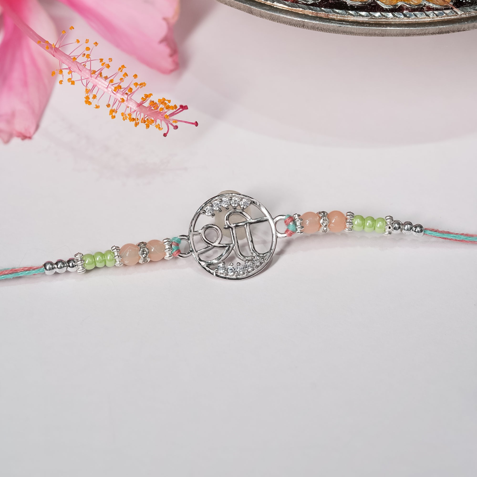 Silver rakhi with Shree symbol on saffron-red thread, displayed on minimal background.

