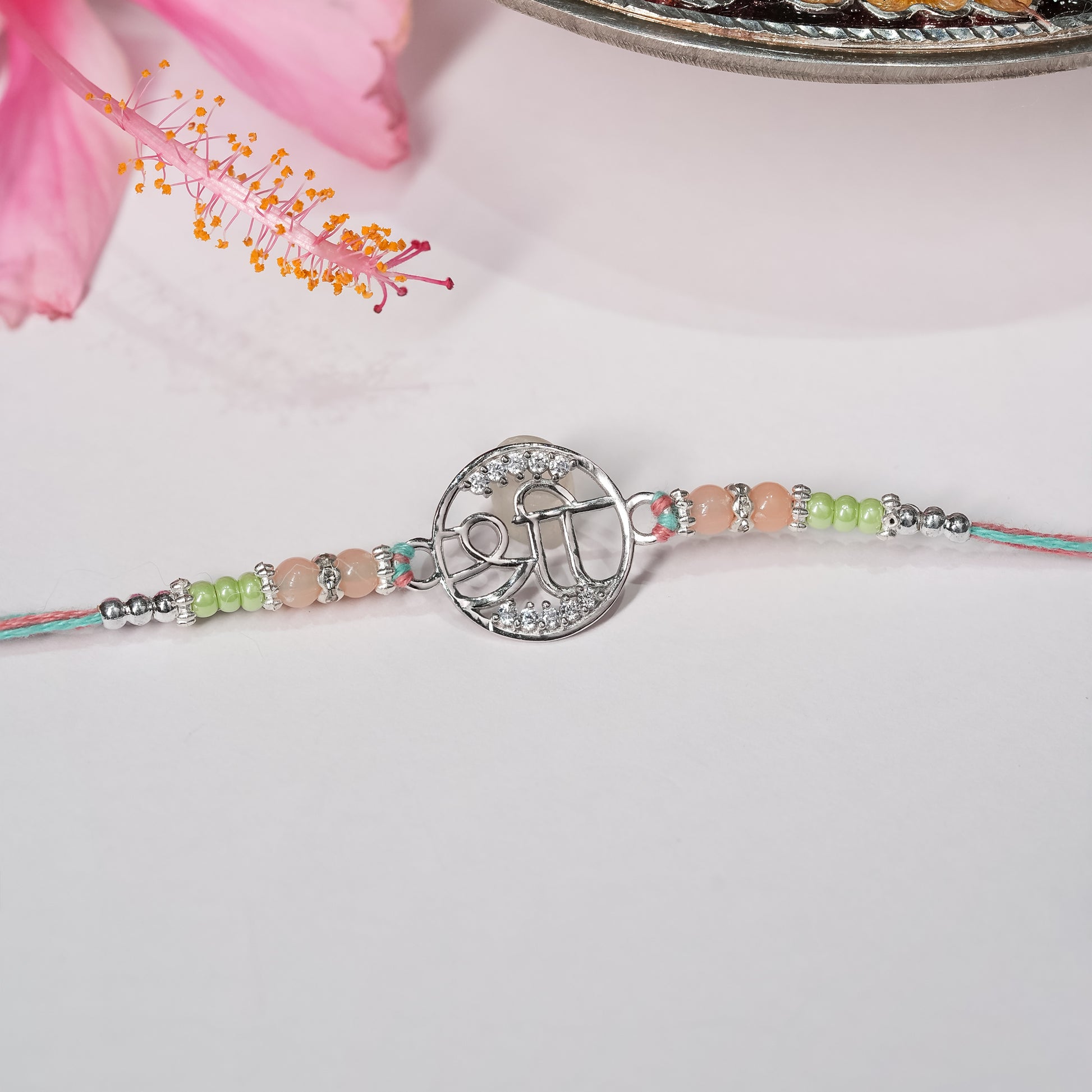 Silver rakhi with Shree symbol on saffron-red thread, displayed on minimal background.

